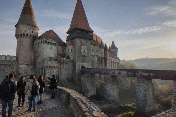 A guide and a small group beside the pointed towers and steep roofs of a vast gothic castle on a rocky foundation, Castelul Corvinilor