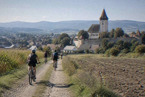 A small group on bikes in helmets riding a dirt track between fields toward a village with a fortified church tower, Cisnădie