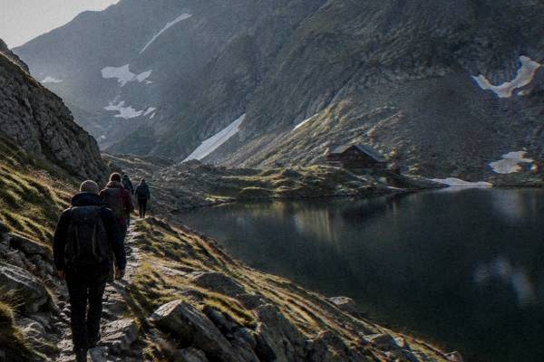 Hikers with daypacks on a stony mountain path beside a still dark lake under steep rock walls, seen from behind, Lacul Bâlea