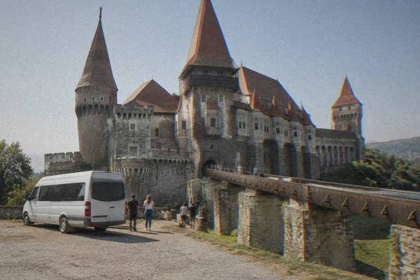 A plain minibus parked beside a long wooden bridge leading to a huge castle with many steep-roofed towers, visitors beside it, Castelul Corvinilor