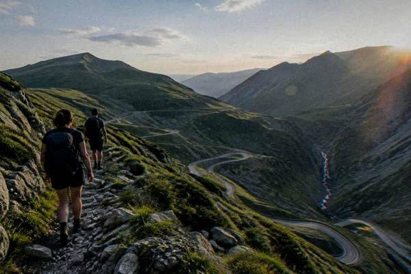 Two hikers with daypacks on a rocky ridge path above scree slopes under a wide sky, seen from behind, Transfăgărășan