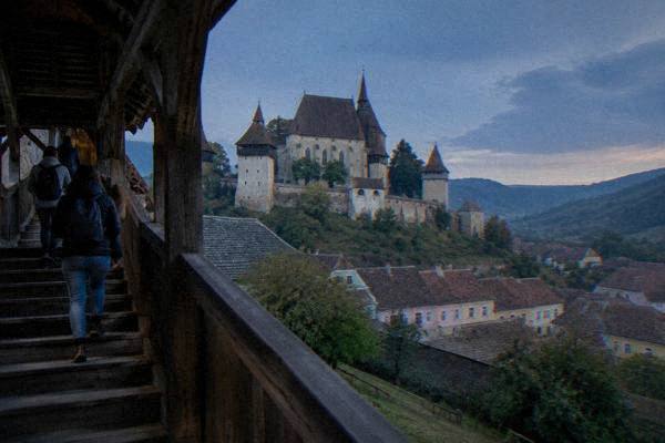 A small group on a covered wooden stair climbing toward a hilltop fortified church inside thick walls, Biertan