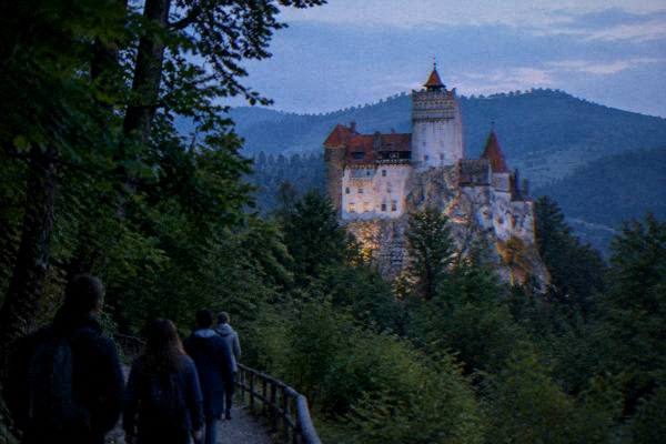 A small group on a path through green forest looking up at a white-walled castle with steep orange roofs on a rocky outcrop, Castelul Bran