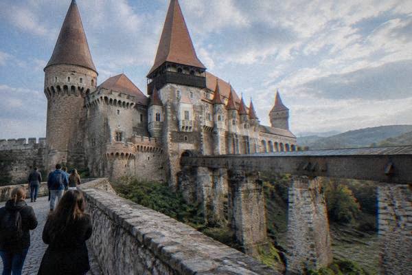 Visitors on a stone bridge over a dry moat looking up at the many steep-roofed towers of a gothic castle, Castelul Corvinilor