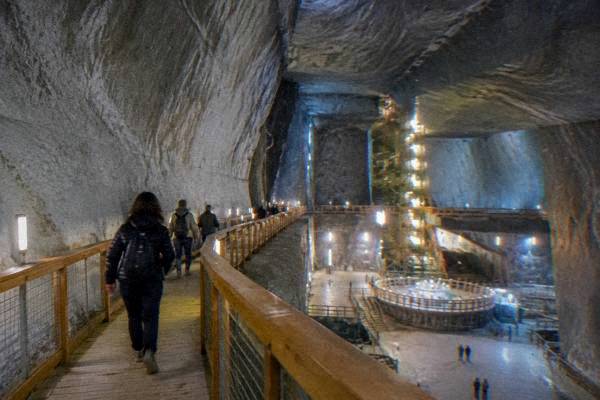 Visitors on a wooden walkway inside a vast underground salt mine hall with smooth pale walls and soft lamps, Salina Turda
