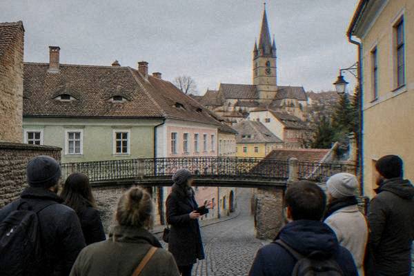 A guide telling a story to a loose circle of visitors beside a short low iron bridge over a cobbled lane in morning light, Podul Minciunilor
