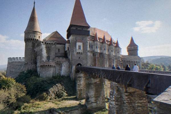 A small group of day visitors on a long covered wooden bridge leading to an enormous gothic castle with pointed towers, Castelul Corvinilor