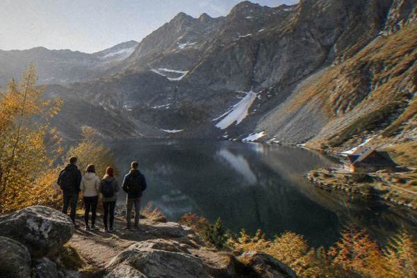 A small group of visitors standing at a viewpoint beside a dark glacial lake under steep bare rock walls, seen from behind, Lacul Bâlea