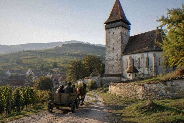 A horse-drawn farm cart on a dirt lane beside a stone fortified church with a tall tower, a few visitors on board, Valea Viilor
