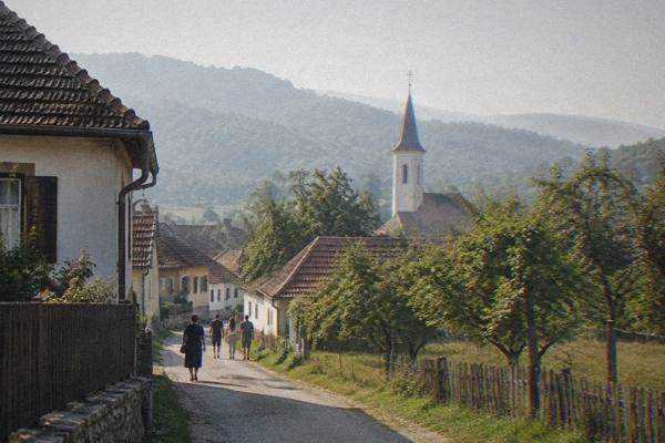 A small group strolling along a village lane of low traditional houses past orchards, a slim church tower behind, Sibiel