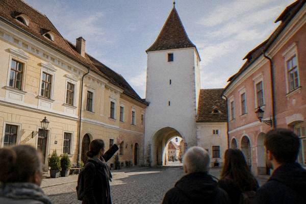 A guide gesturing up at a tall plain white tower over a vaulted passage between two squares while a small group listens, Turnul Sfatului