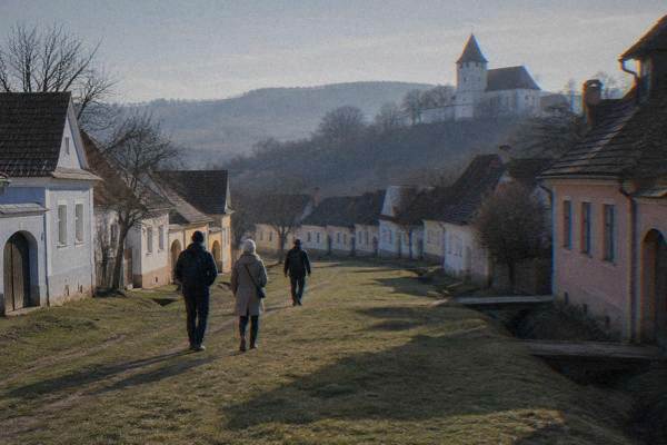A couple and a guide walking down the wide grassy street of a tiny village of low pastel houses with gabled tiled roofs, Viscri