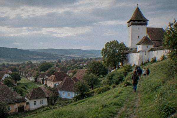 A small group on a hillside path beside a whitewashed fortified church with a plain tower, a village of tiled roofs below, Viscri