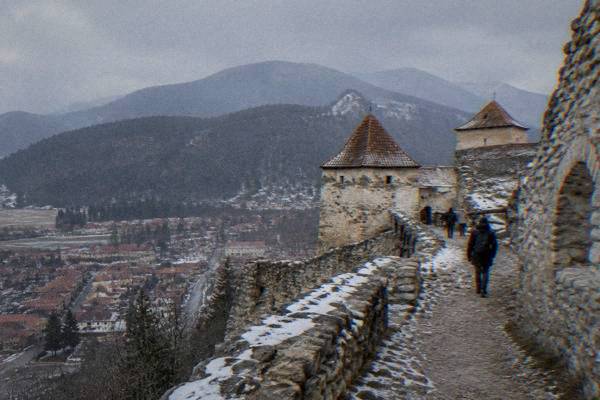A few visitors on a stony path inside the pale stone walls of a hilltop peasant fortress, red tiled roofs of a town far below, Râșnov