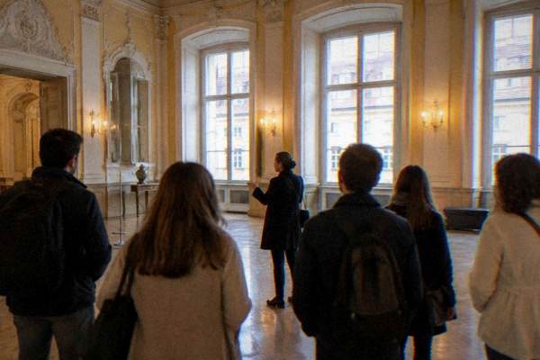 A small group in a bright baroque hall with tall windows and a polished floor listening to a guide, seen from behind, Muzeul Național Brukenthal