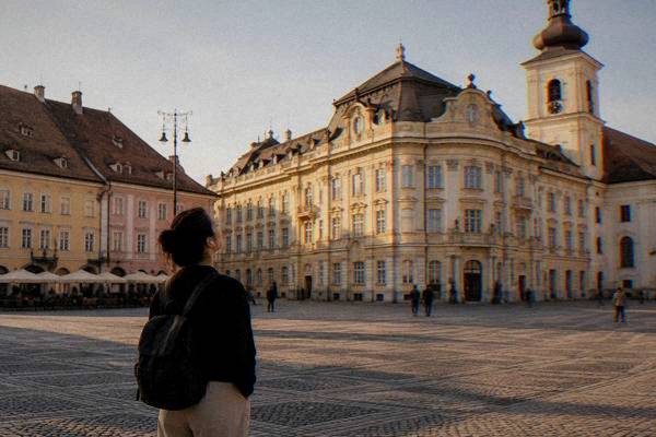 A lone visitor with earphones in, pausing in a very large cobbled square to look up at a pastel baroque palace, seen from behind, Piața Mare