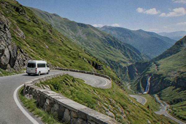 A plain white minibus on a narrow mountain road with sweeping hairpin bends across bare green slopes of high mountains, Transfăgărășan