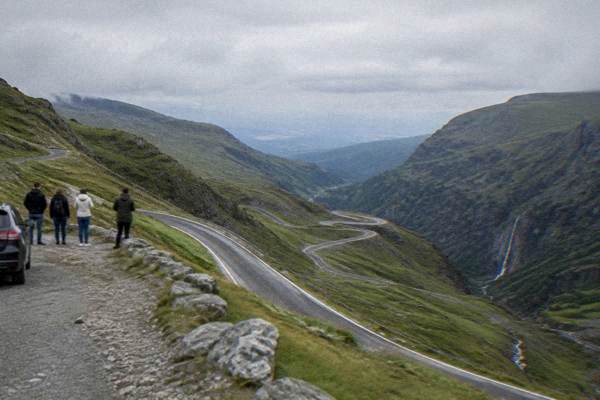 A narrow asphalt road in sweeping hairpin bends across bare high green slopes, a small group of visitors at a lay-by looking out, Transfăgărășan