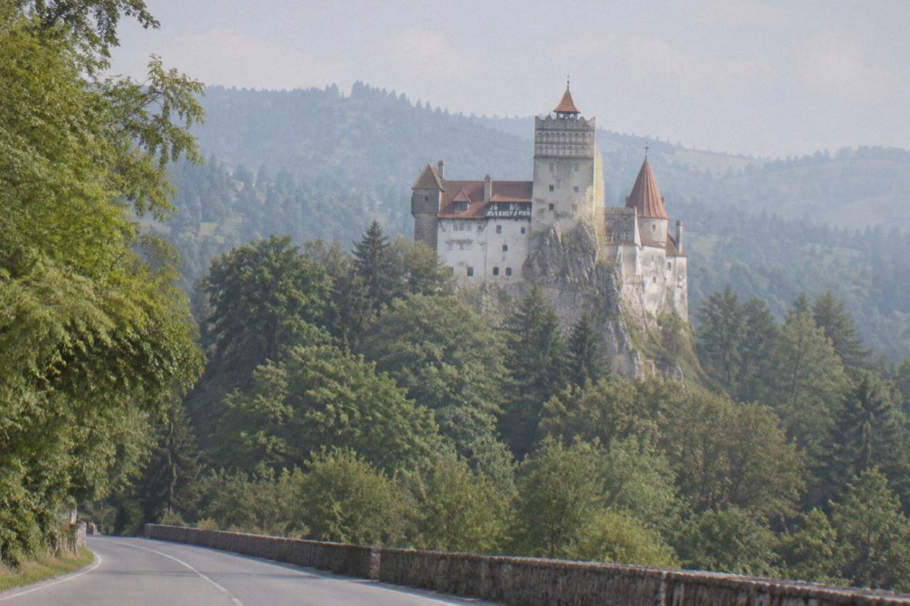 Bran Castle seen from the road below, with its white towers and red-tiled roofs against a wooded hillside