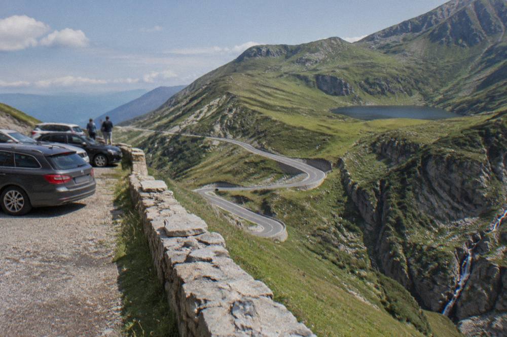 Cars parked at a viewpoint on a high mountain road with a glacial lake in the distance