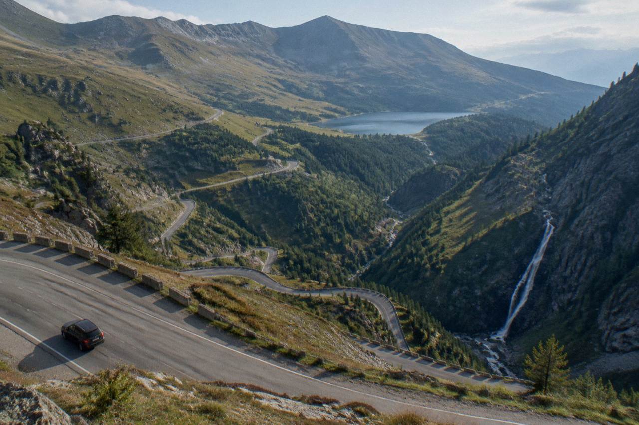 A winding mountain road climbing forested slopes toward a glacial lake high in the Făgăraș mountains