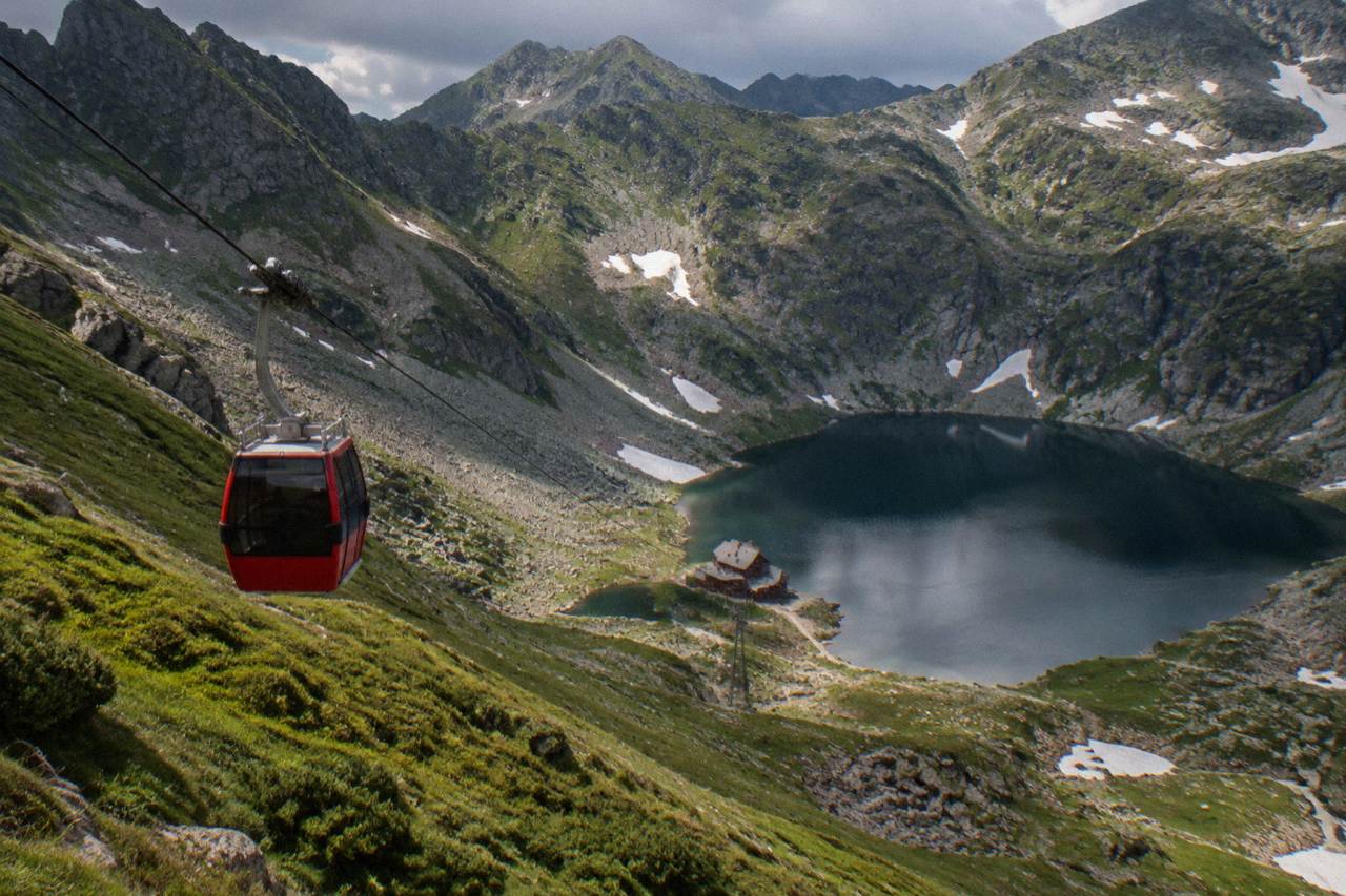 A cable car cabin travelling above a green mountain slope toward a glacial lake ringed by peaks