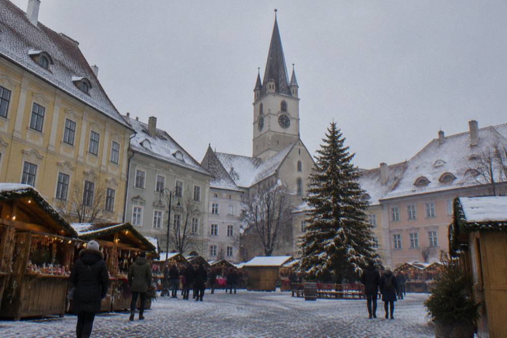 A winter market square in Sibiu with wooden stalls, a lit Christmas tree and snow on the rooftops