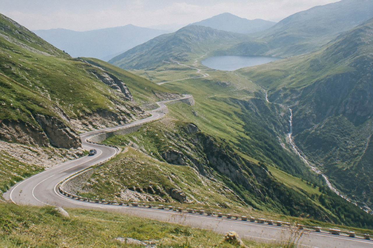 A winding mountain road climbing through green peaks toward a lake high in the Făgăraș mountains
