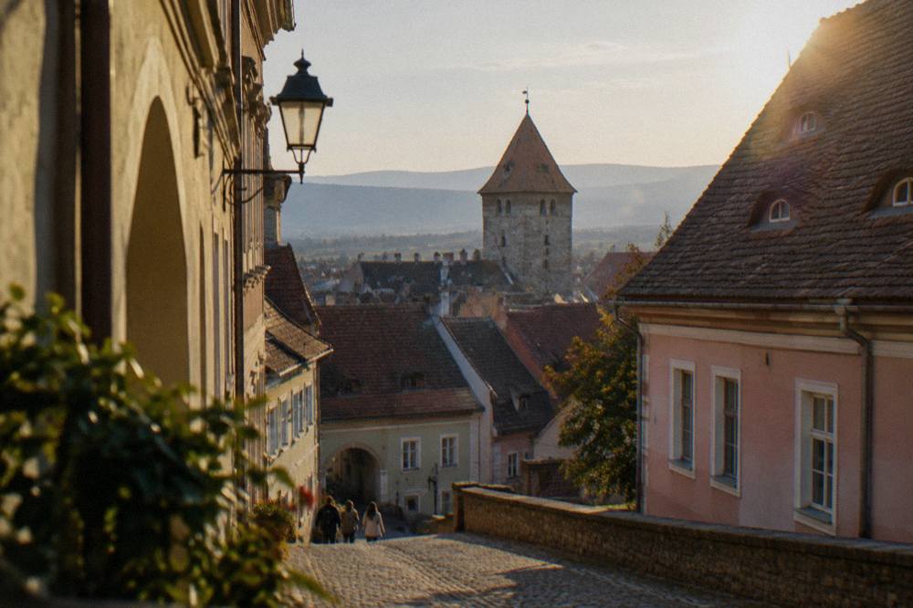 A stone tower with a tiled roof standing above red rooftops on a downhill street in Sibiu