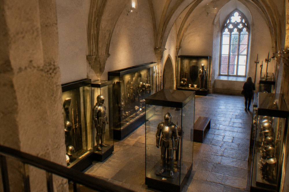 Display cases of medieval weapons and armour under a vaulted ceiling