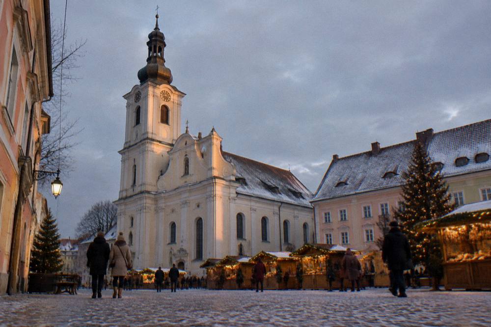 A historic square at dusk with Christmas market lights and a church tower in the background