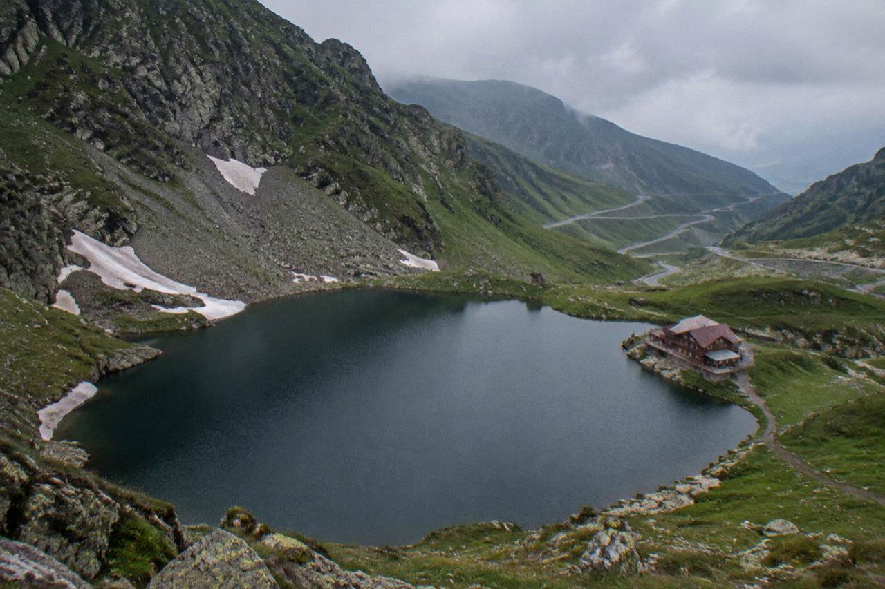 A glacial lake ringed by steep green slopes under a cloudy sky, with a road switchbacking down the mountainside behind it