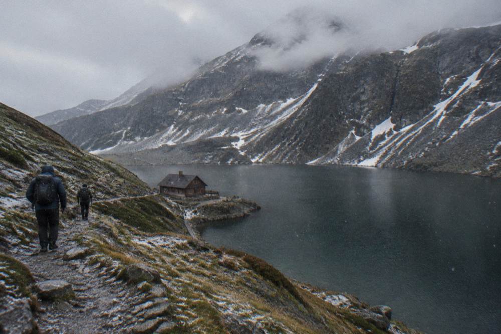 Hikers on a rocky path above a mountain lake, ridgeline clouds moving across the peaks behind them