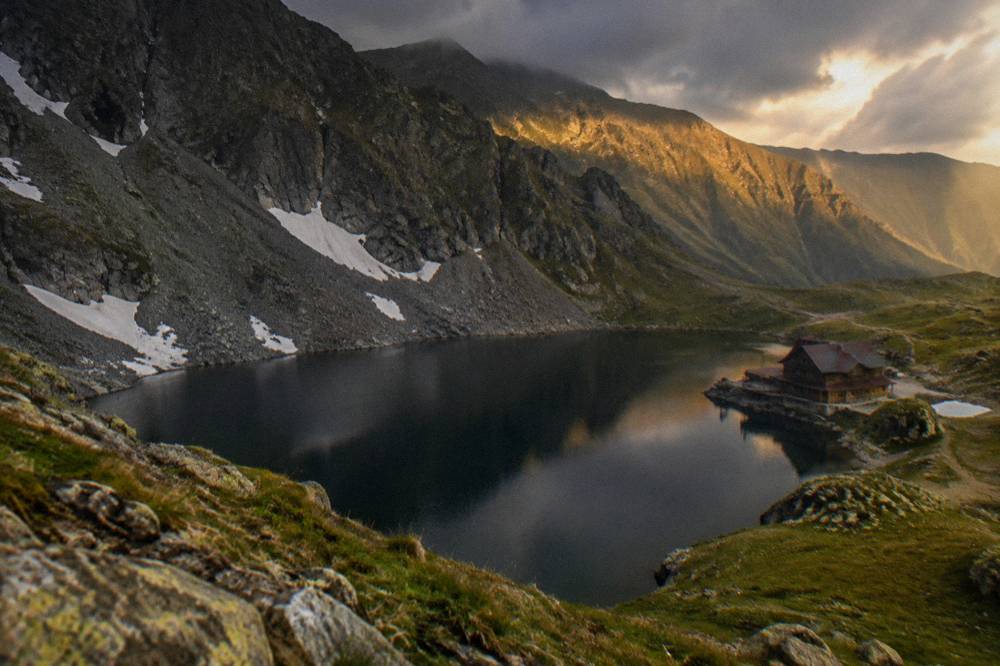 Dark clouds passing over a mountain crest with a lake below, shafts of light on the far ridge