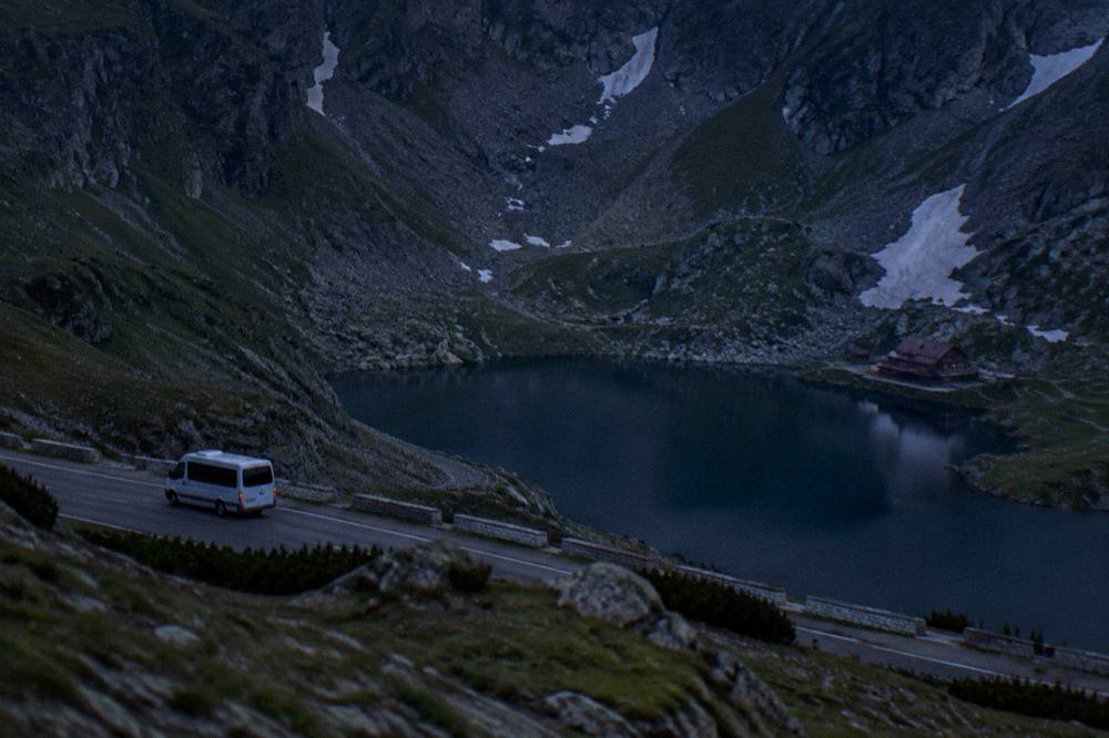 A minibus on a mountain road above a valley, the crest and a lake visible in the distance