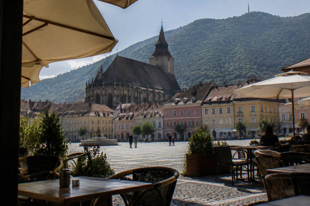 An outdoor cafe terrace on the edge of Council Square in Brașov with mountain ridges visible above the rooftops