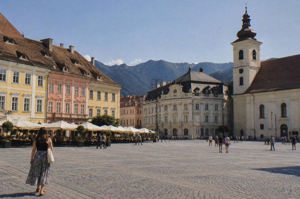 The main square of Brașov in late afternoon light with cafe umbrellas and mountain ridges behind the rooftops