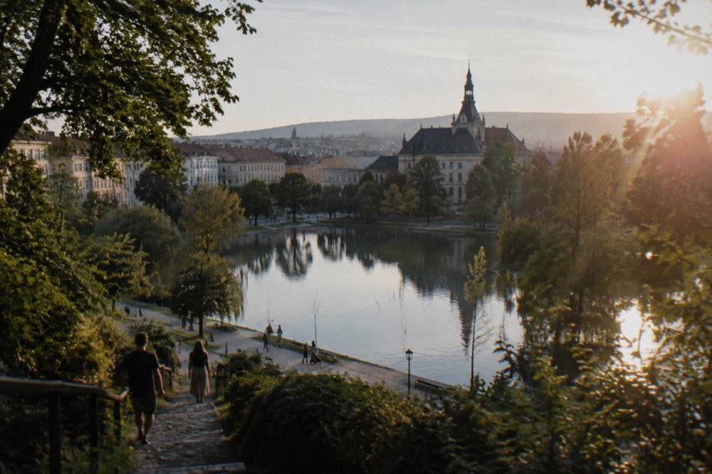 People strolling beside a lake in a city park with a historic building among trees