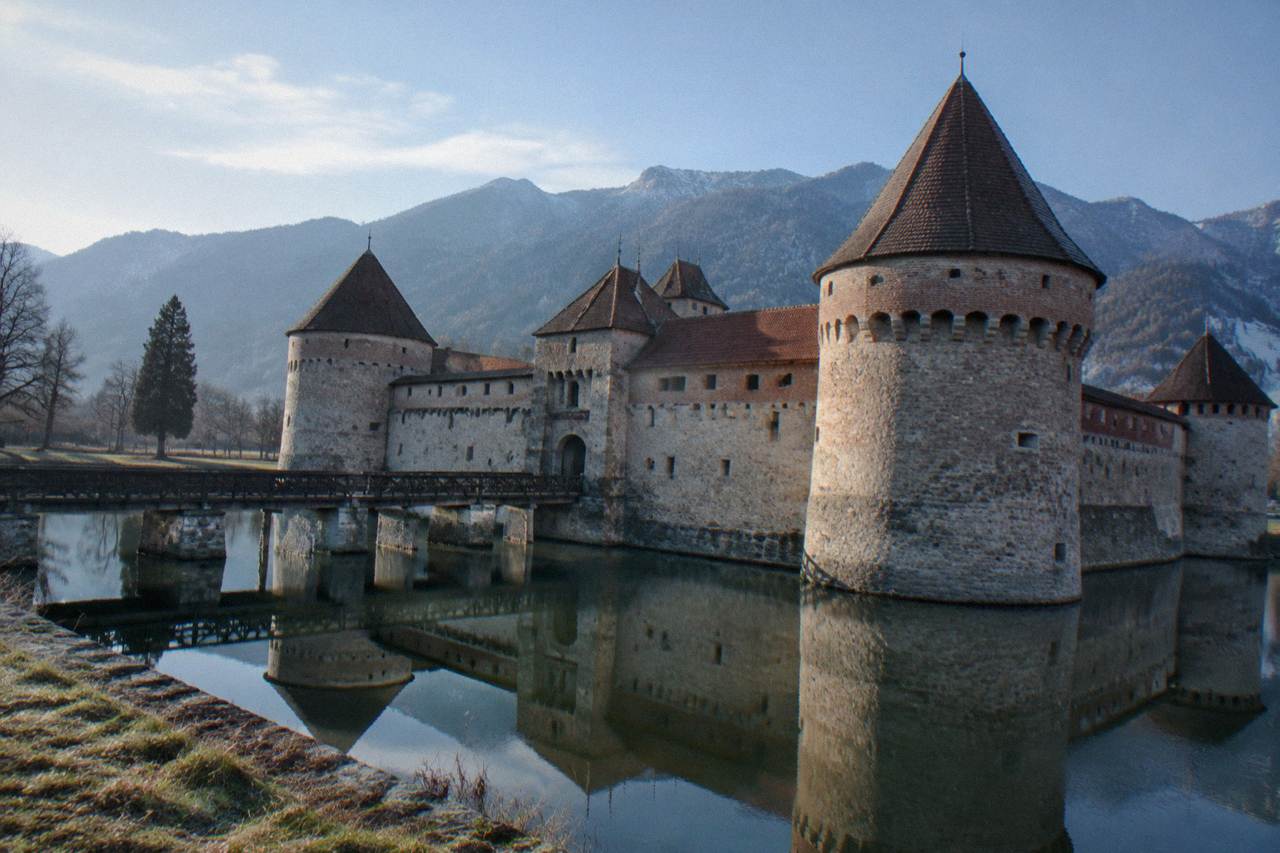 A moated fortress with round corner towers reflected in still water, tree covered mountains behind it