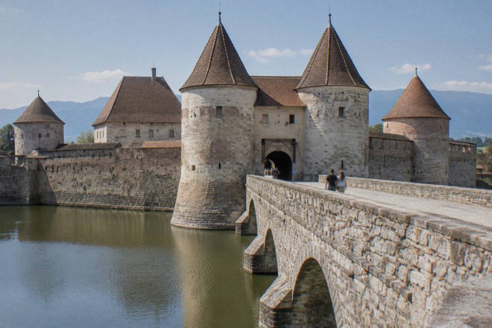 A stone bridge crossing a water moat toward a fortified gatehouse with twin towers
