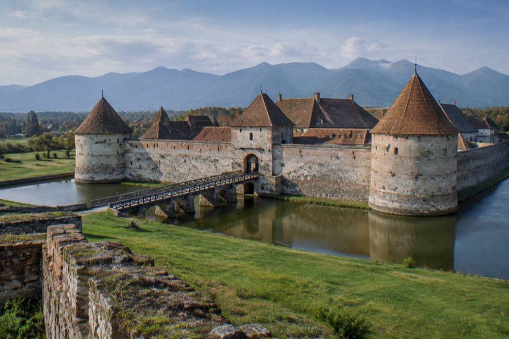 A fortified castle wall seen across a wide grassy field with mountain peaks in the far distance