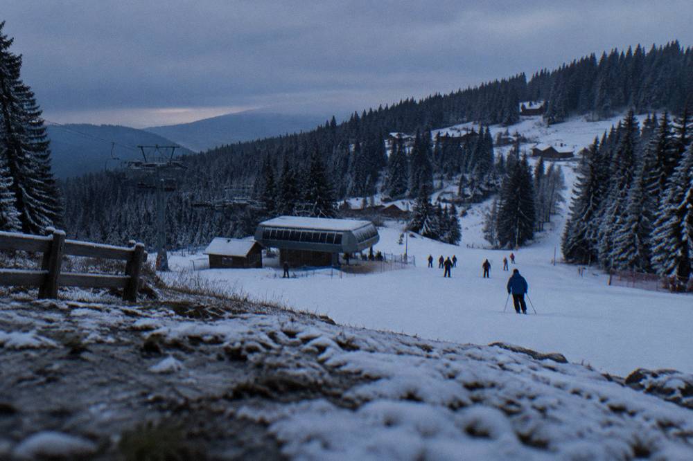 Skiers on a modest piste beside a lift station with snow-covered spruces
