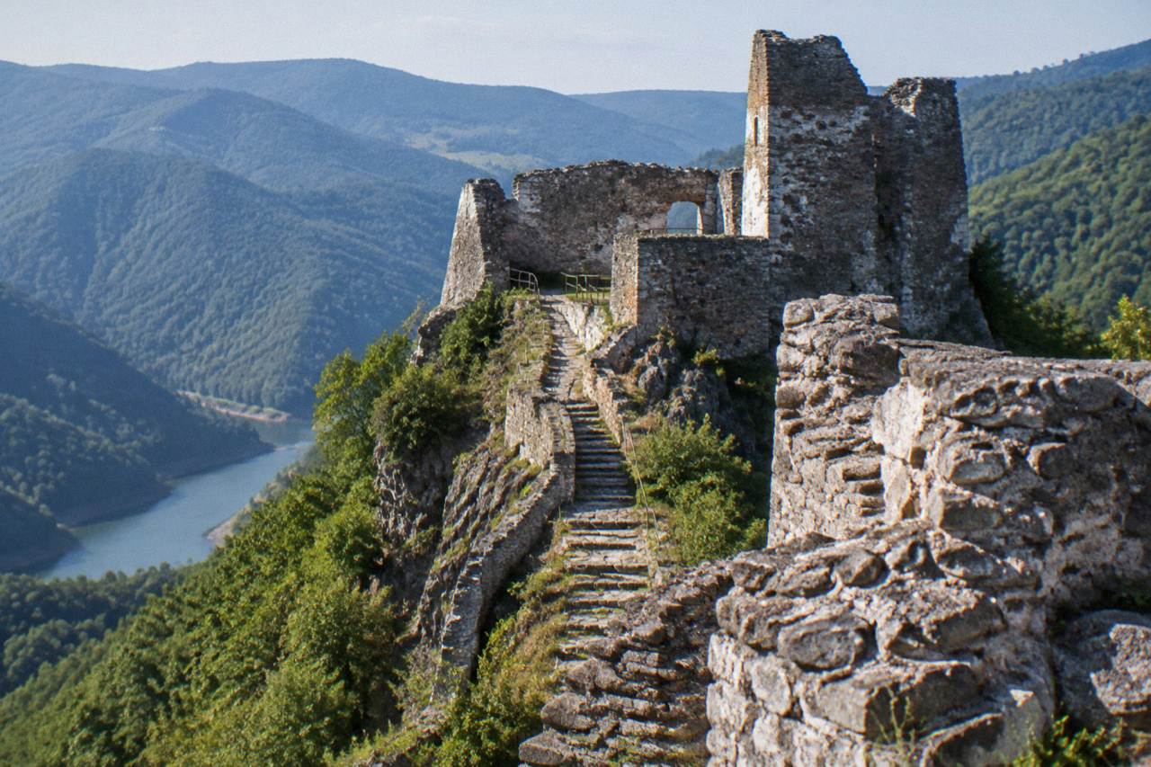 A ruined stone fortress on a forested rocky ridge above a valley in the Transylvanian mountains
