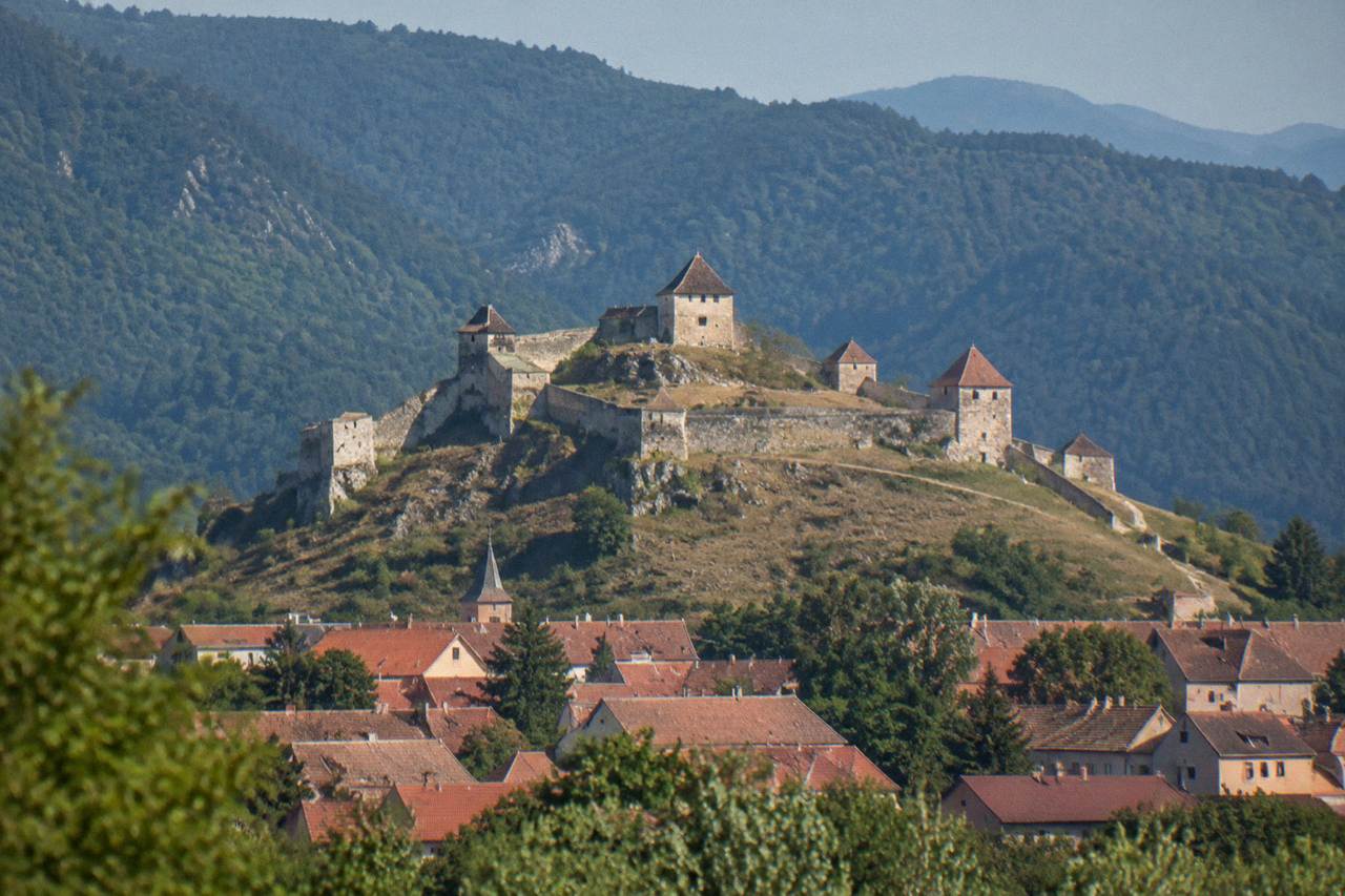 A walled hilltop citadel with towers rising above a small town and green hills