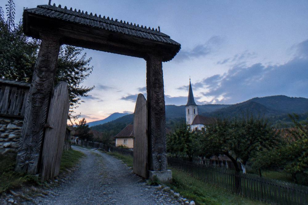 A wooden village gate with carved posts standing open onto a gravel lane