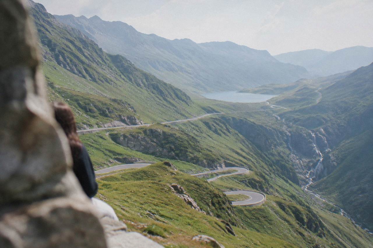 A winding mountain road climbing through green peaks toward a glacial lake