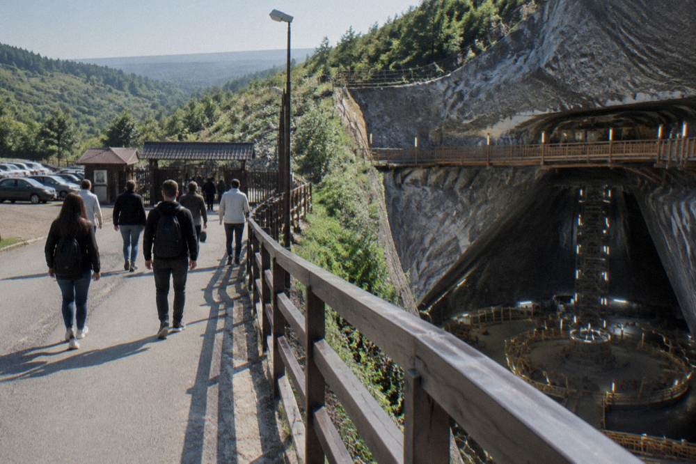 A car park entrance at a hillside site with visitors walking toward a gate in late morning light