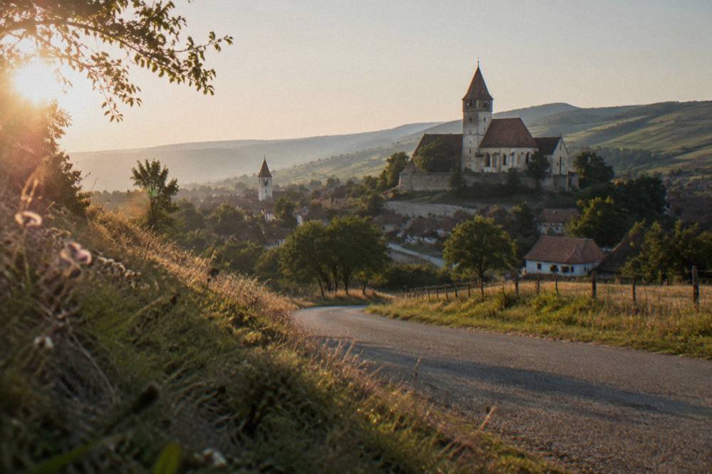 A country road winding through hay meadows and low hills toward a village with a church tower