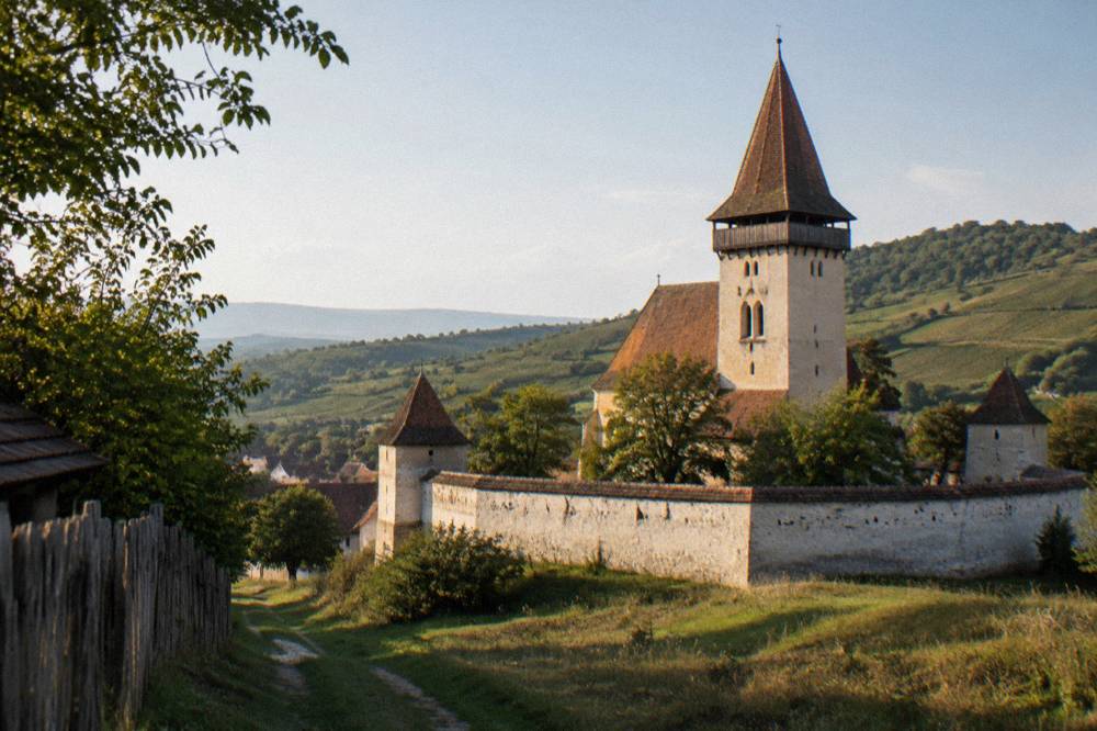 The walled enclosure of a village church fortress seen from a grassy lane in late afternoon light