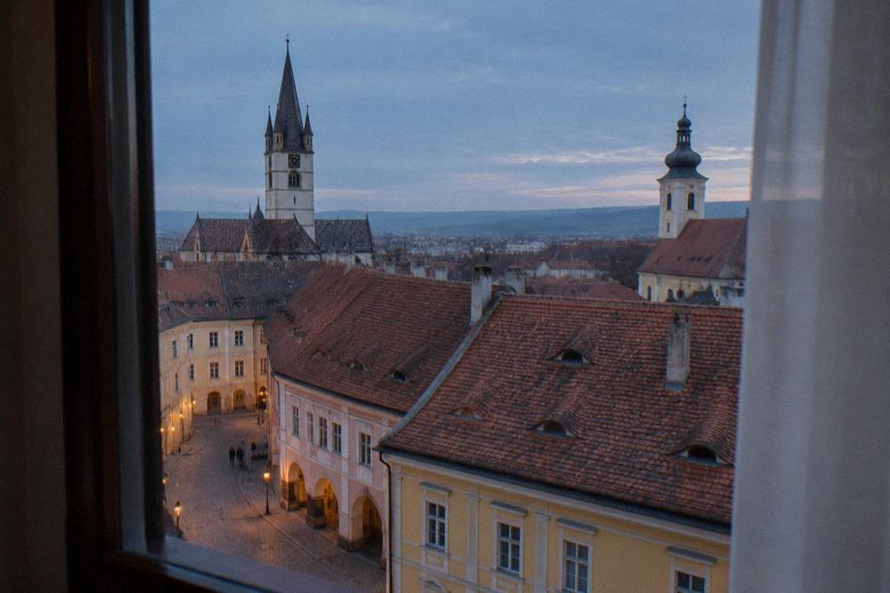 A hotel room window overlooking red tiled roofs and church towers in Sibiu
